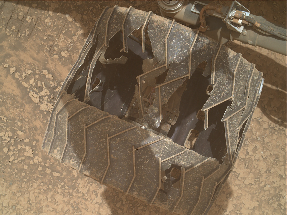 A wheel of the Curiosity rover on the Martian surface. The outer casing looks extensively damaged, with a lot of the inside of the wheel clearly visible.